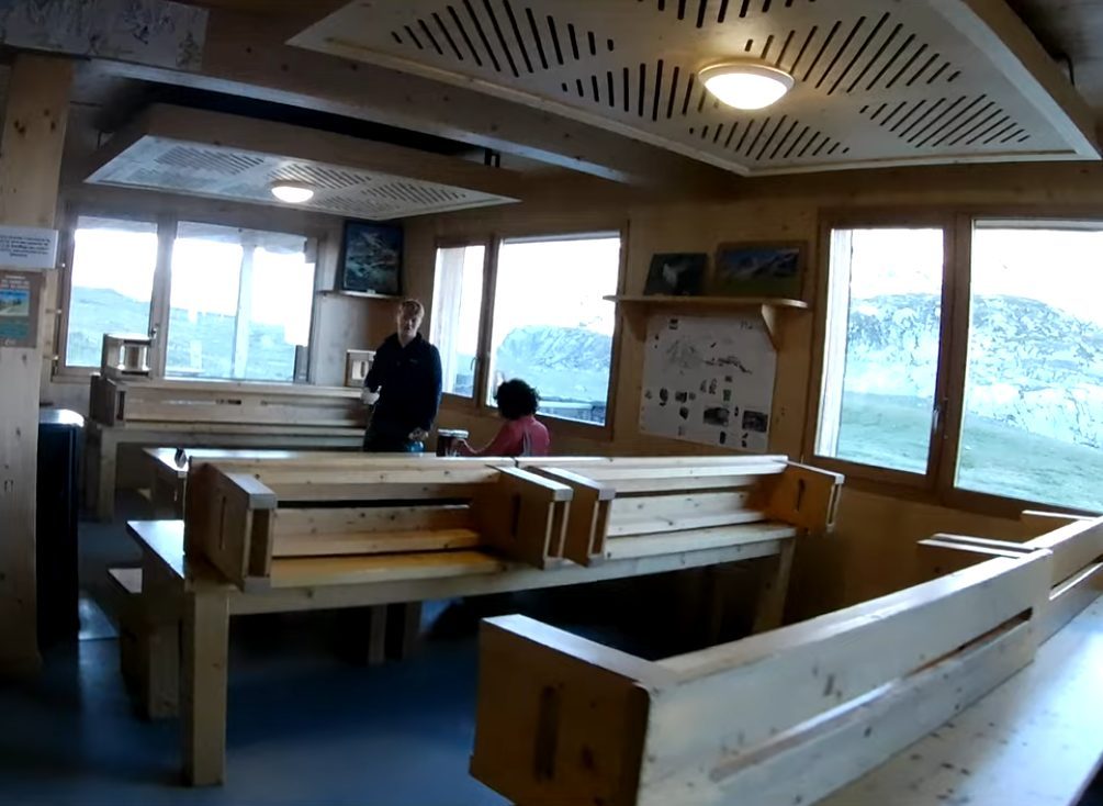 The long shared tables of a hut dining room, with snow and rock through the windows.