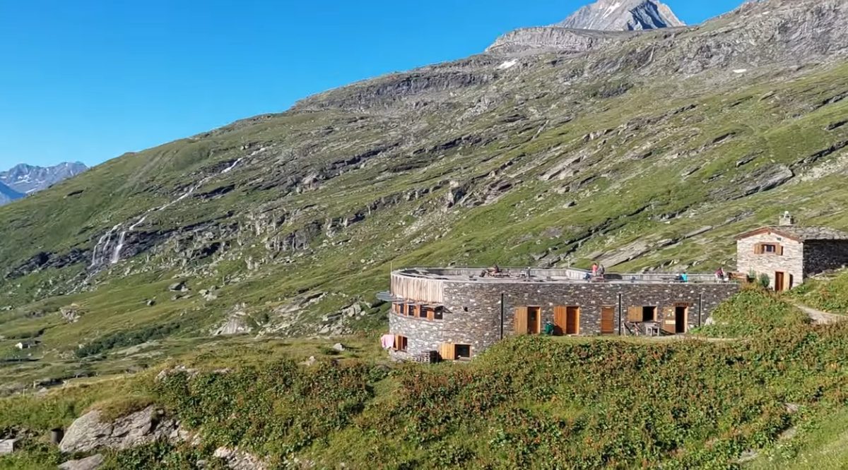 A stone-built refuge on an alpine hillside, its terrace above the valley and rock walls behind.