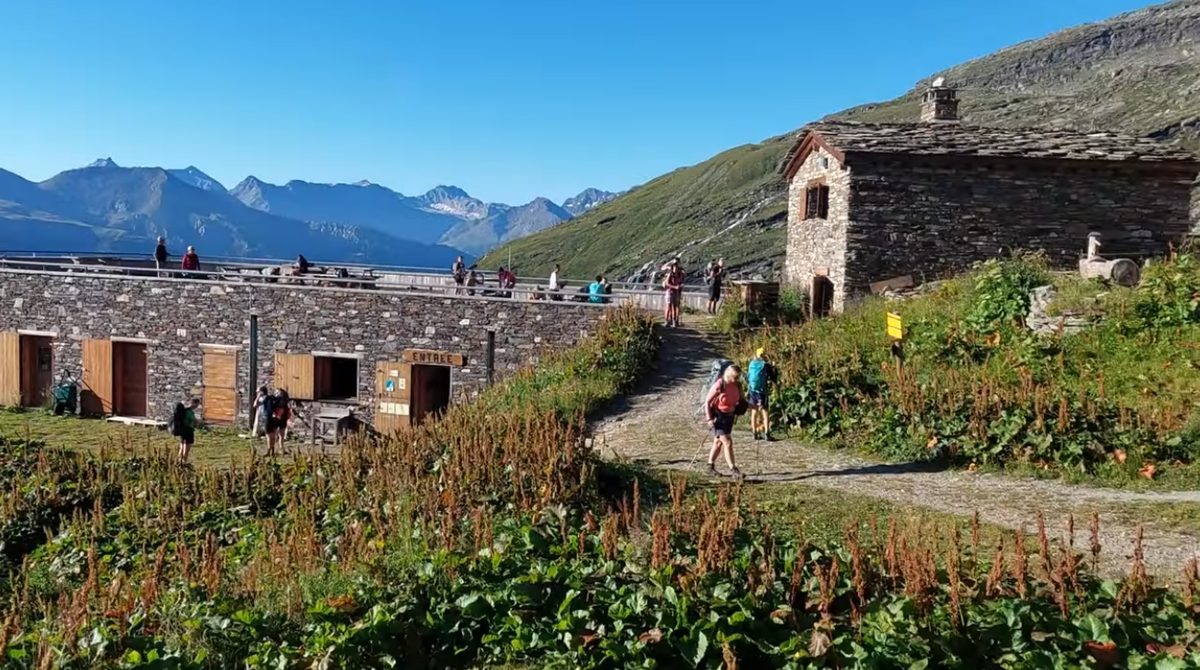 Walkers setting out from a refuge on a summer morning, its terrace already lined with people.