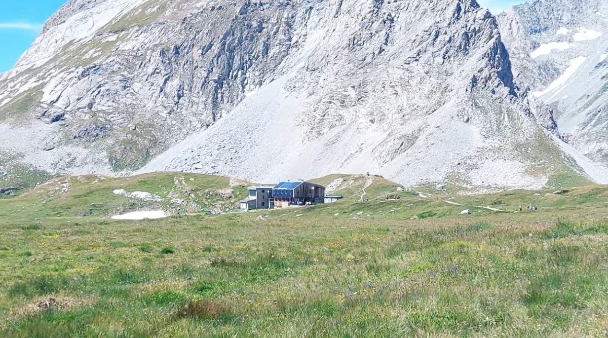 One hut alone on a broad meadow beneath a great rock face, with a few walkers on the path.